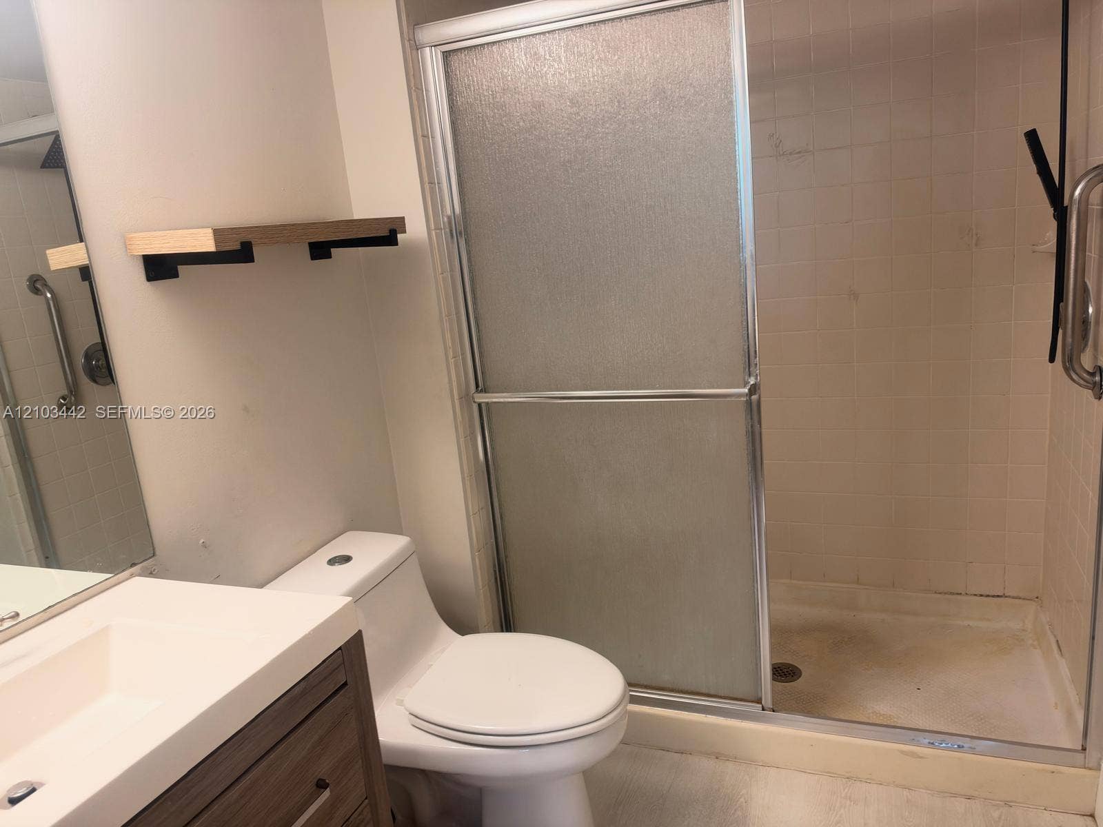 8721 Wiles Road, #303, Coral Springs, FL 33067 Bathroom featuring a vanity with a white composite countertop, a wood-finish cabinet, a walk-in shower with a frosted glass sliding door, and wall-mounted shelving