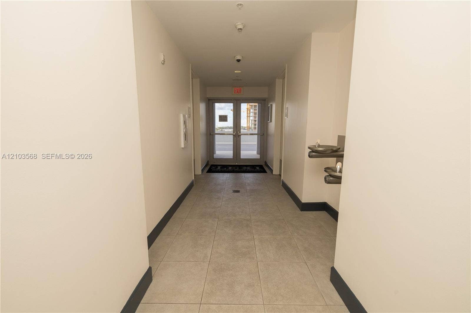7901 Hispanola Avenue, #801, North Bay Village, FL 33141 Interior hallway featuring neutral walls, large-format tile flooring, double glass-paneled doors, and wall-mounted metal water fountain station