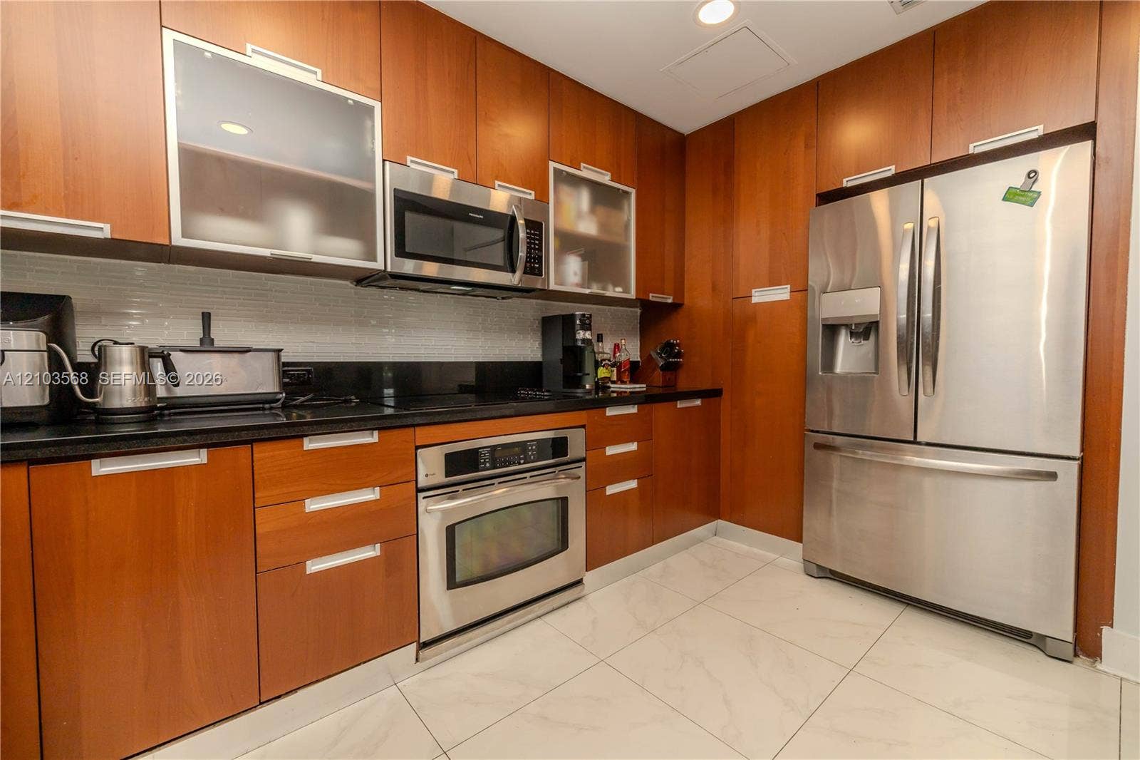 7901 Hispanola Avenue, #801, North Bay Village, FL 33141 Kitchen featuring wood-finish cabinetry, stainless steel appliances, dark stone-style countertops, white subway tile backsplash, and large-format tile flooring