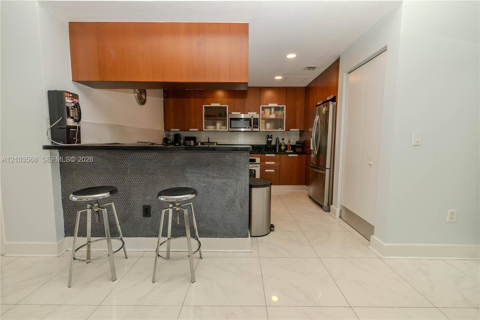 7901 Hispanola Avenue, #801, North Bay Village, FL 33141 Kitchen featuring wood-finish cabinetry, a dark stone countertop, a textured breakfast bar, stainless steel appliances, and large-format tile flooring