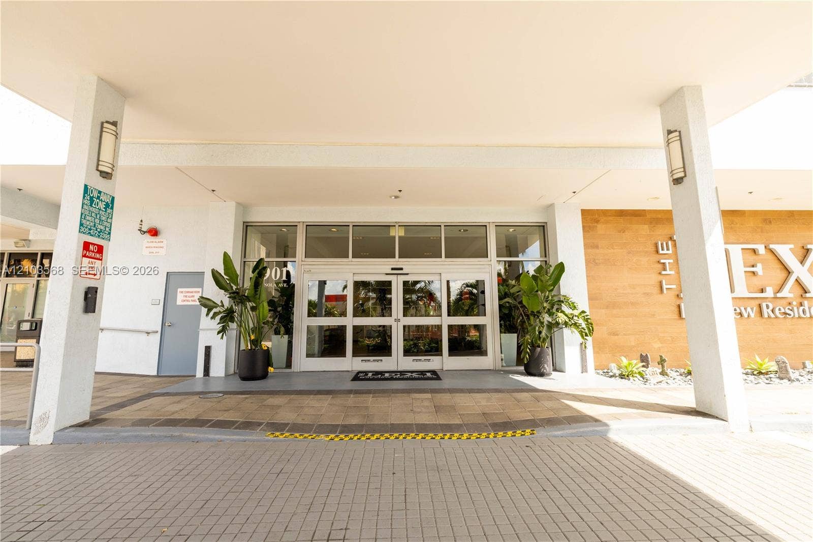 7901 Hispanola Avenue, #801, North Bay Village, FL 33141 Covered building entrance featuring a stone-paved driveway, multi-pane glass sliding doors, wall-mounted exterior sconces, and wood-finish paneling