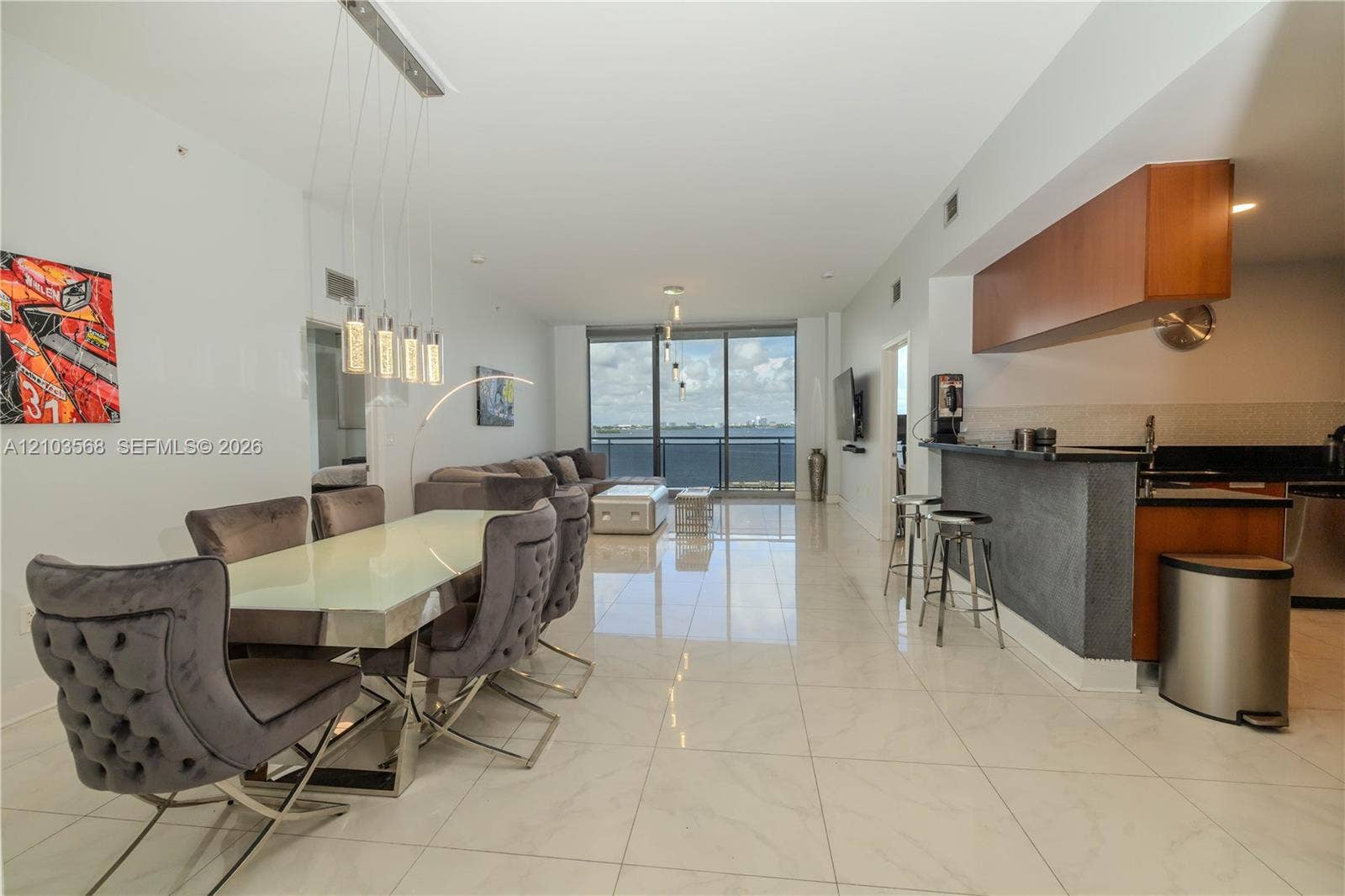7901 Hispanola Avenue, #801, North Bay Village, FL 33141 Open-concept interior featuring large-format tile flooring, a kitchen breakfast bar with stone-texture base, wood-finish upper cabinetry, a five-light linear pendant fixture, and floor-to-ceiling glass panel windows