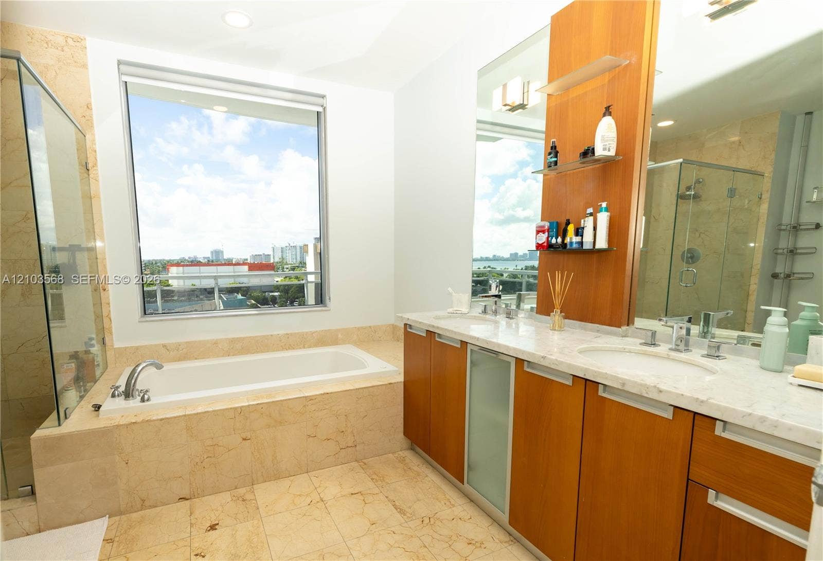 7901 Hispanola Avenue, #801, North Bay Village, FL 33141 Bathroom featuring a deep soaking tub with a stone surround, dual-sink vanity with light-toned stone countertops, natural wood-finish cabinetry, a large frameless glass-enclosed shower, and expansive windows