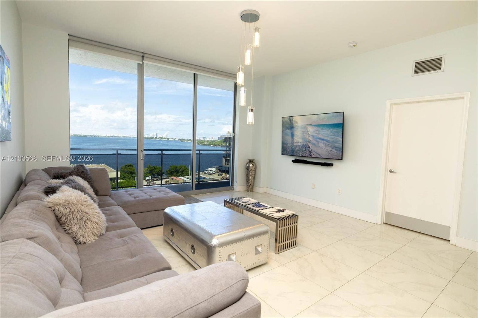 7901 Hispanola Avenue, #801, North Bay Village, FL 33141 Living space featuring floor-to-ceiling glass sliding doors, stone-finish tile flooring, a tiered multi-pendant light fixture, and a balcony with metal railings