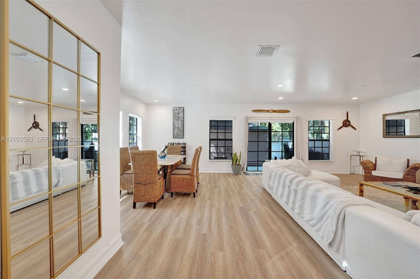 Open-concept interior featuring light wood-finish flooring, grid-patterned floor-to-ceiling wall mirror with brass finish, recessed lighting, and sliding glass doors with black mullions