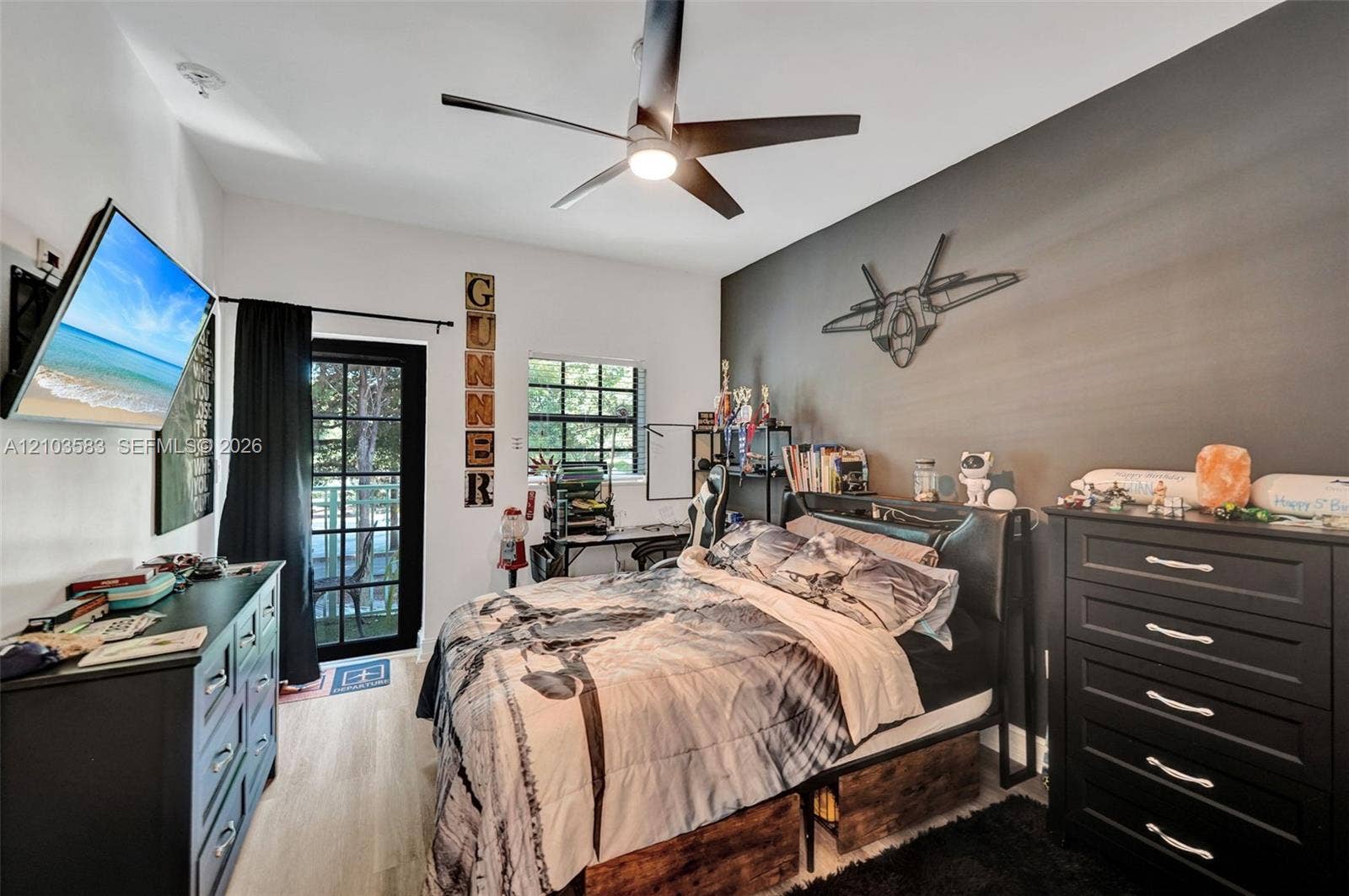 Bedroom featuring light wood-finish flooring, a matte black multi-blade ceiling fan, a dark gray accent wall, and a multi-pane glass-panel exterior door