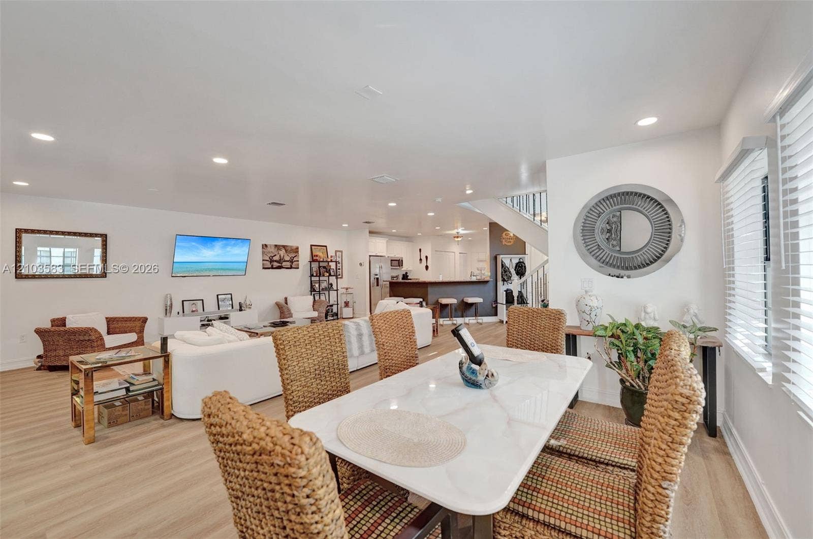 Open-concept interior featuring light-tone wood-finish flooring, recessed lighting, a breakfast bar with stone-finish countertops, and white wall-mounted window blinds