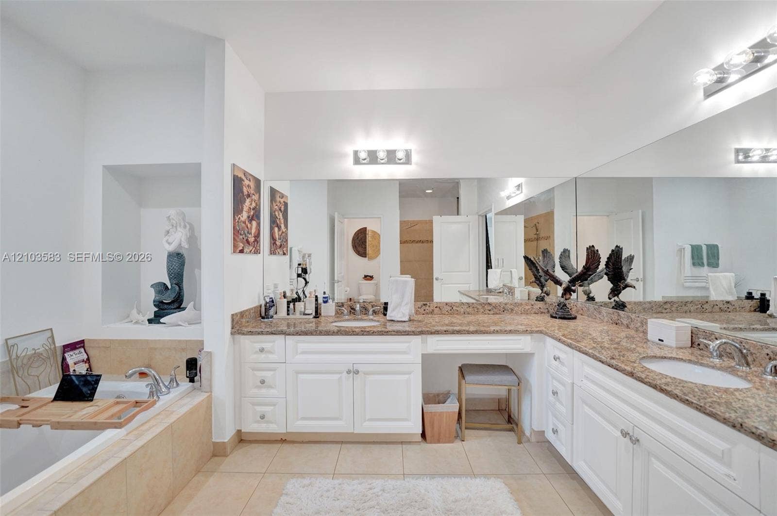 Dual-sink vanity with white cabinetry, stone-finish countertops, built-in makeup vanity, large wall-spanning mirror, and recessed soaking tub with tile surround