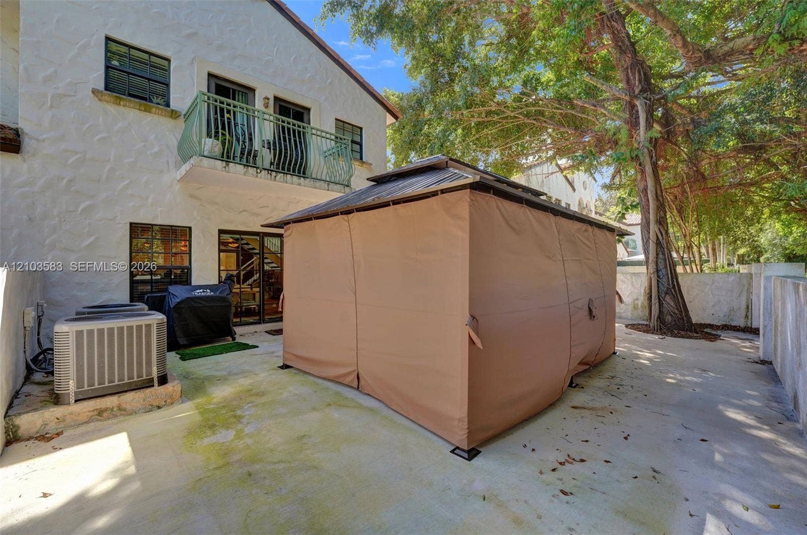 Stucco exterior featuring a second-story balcony with metal railing, concrete patio, and mature trees