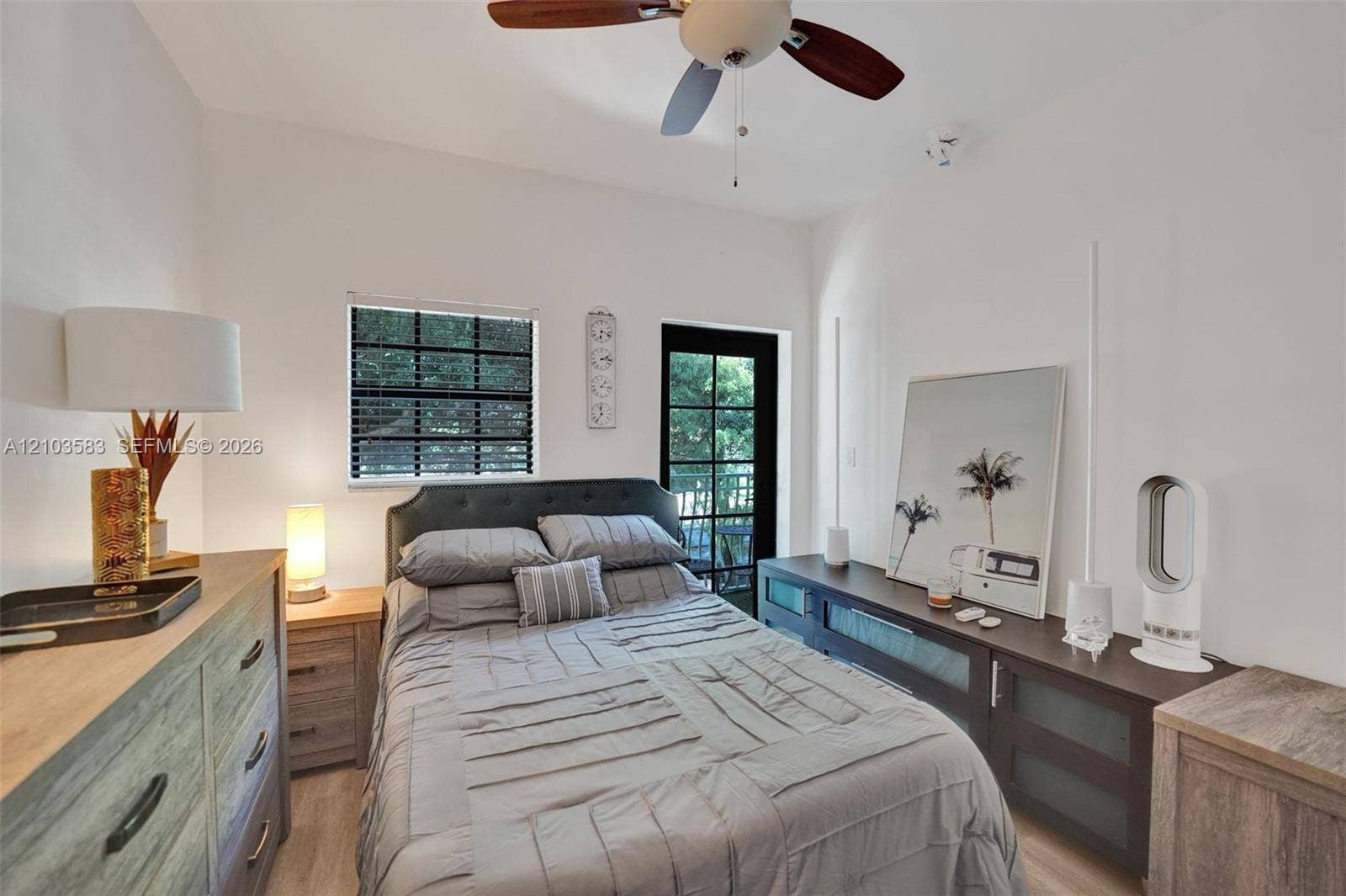 Neutral-toned room featuring a wood-finish floor, a ceiling fan with integrated lighting, a single-pane window with dark-frame blinds, and a glass-paneled exterior door