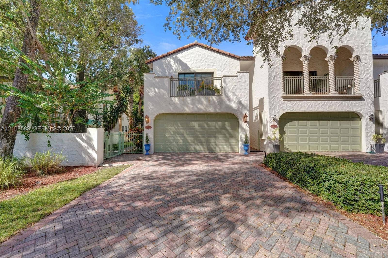 Stucco exterior featuring dual-car garages, a decorative second-story balcony with columns, and a herringbone-patterned paver driveway