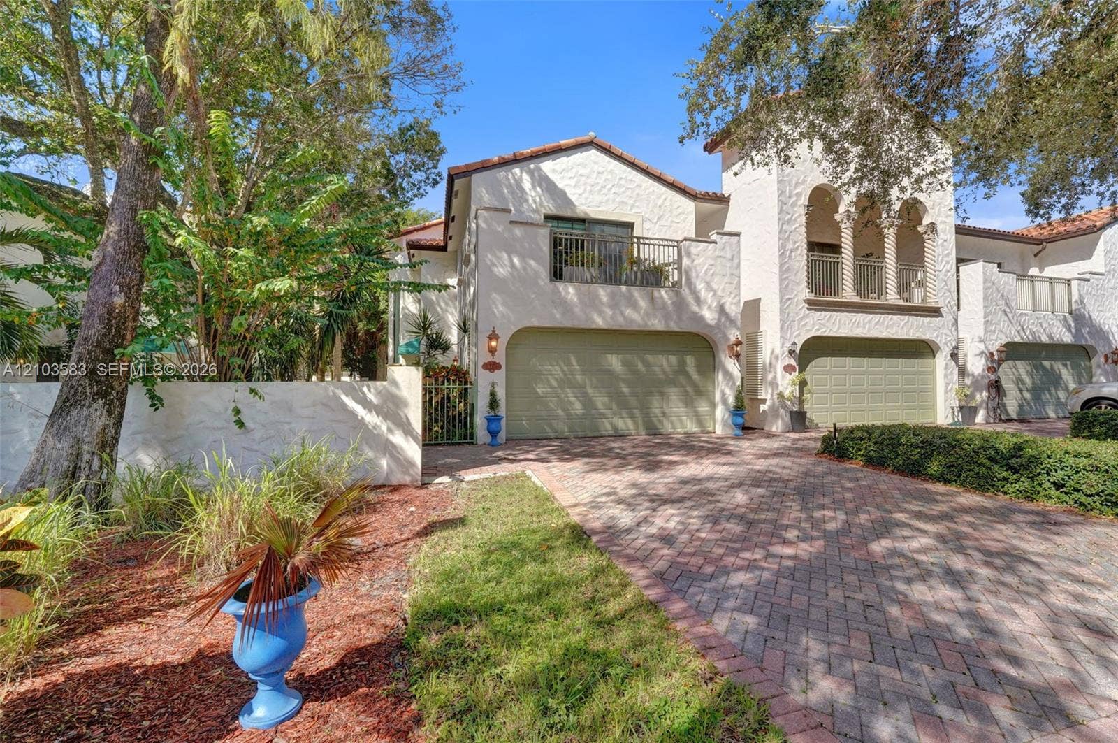 Stucco exterior featuring arched architectural detailing, multiple garage bays, and integrated upper-level balconies with metal railings, set against a brick-paver driveway