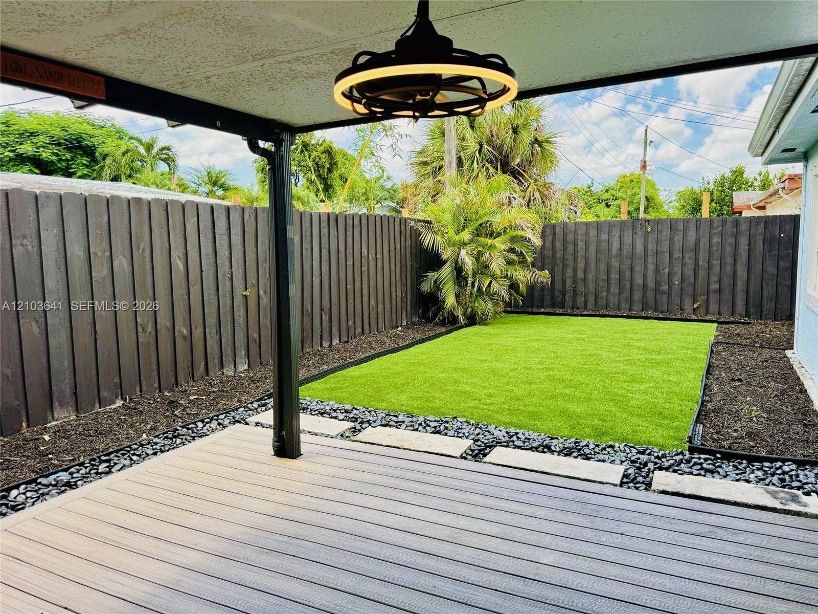 Covered patio featuring composite decking and a modern overhead light fixture, complemented by a fenced backyard with artificial turf, stone hardscaping, and dark mulch garden beds