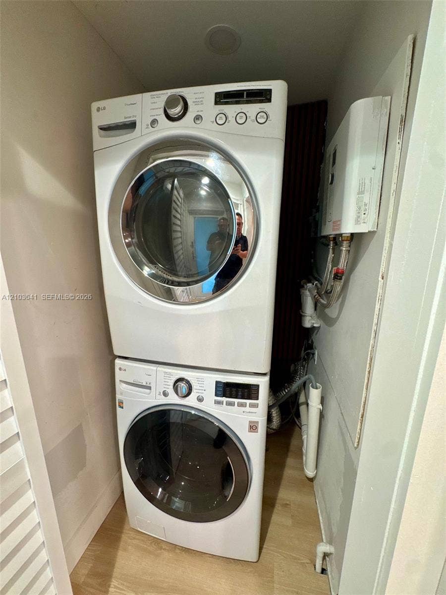 Stacked laundry unit featuring a white front-load washer and dryer with wood-finish flooring and an integrated tankless water heater