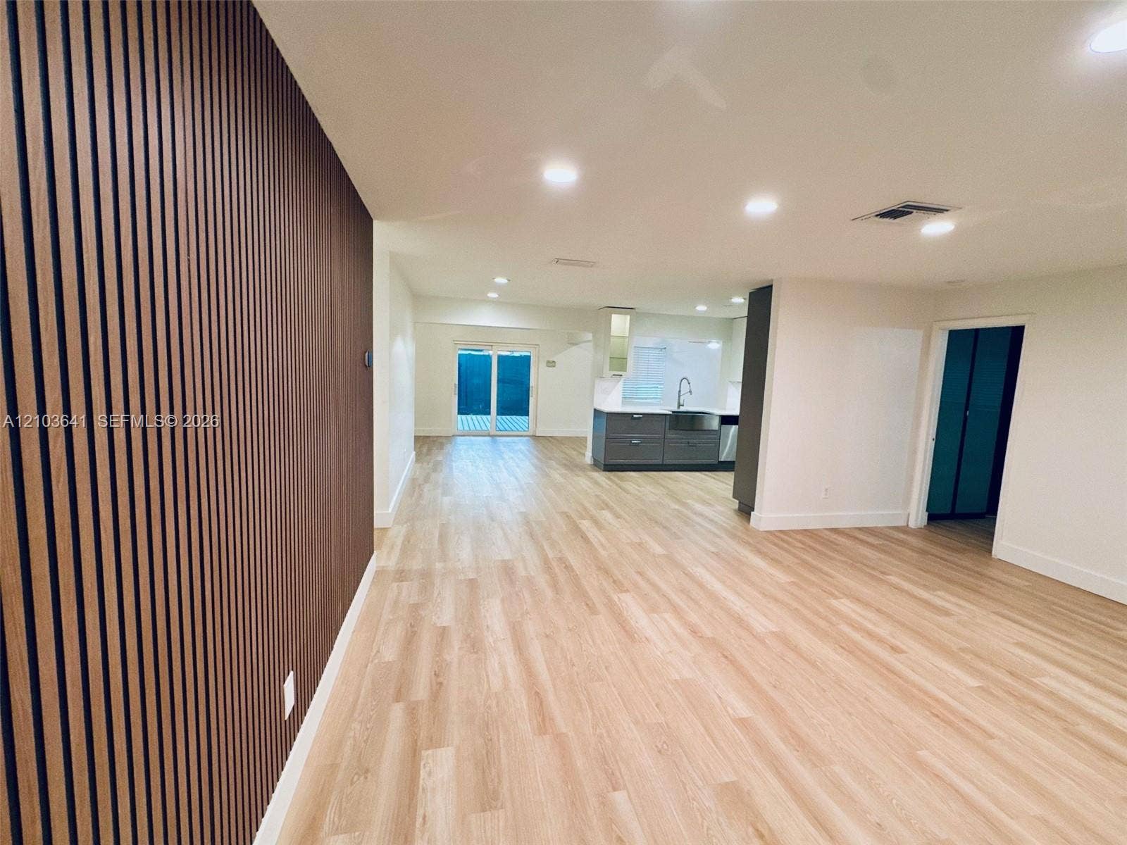 Vertical wood-slat accent wall, wood-finish flooring, recessed lighting, and kitchen area with grey cabinetry and stainless steel apron-front sink