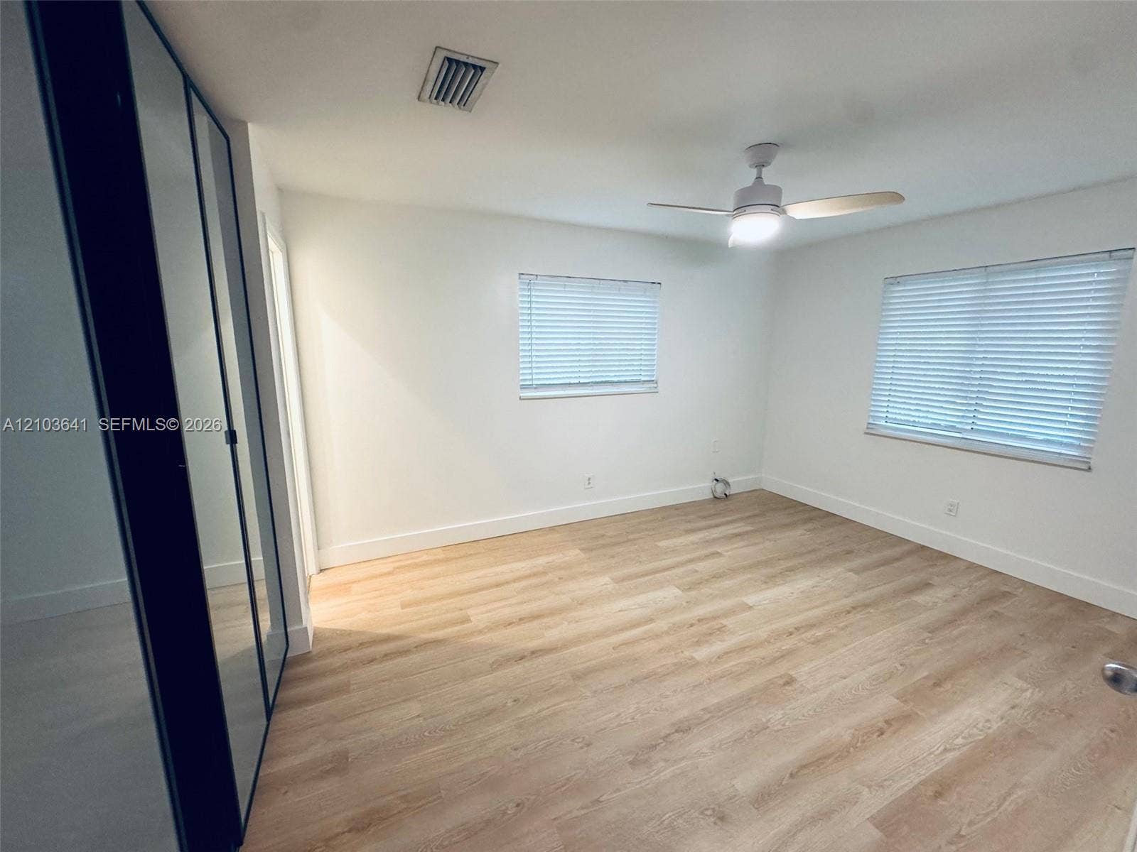 Light-filled room featuring wood-finish flooring, dual windows with blinds, a ceiling fan with integrated lighting, and full-length sliding mirror-paneled closet doors