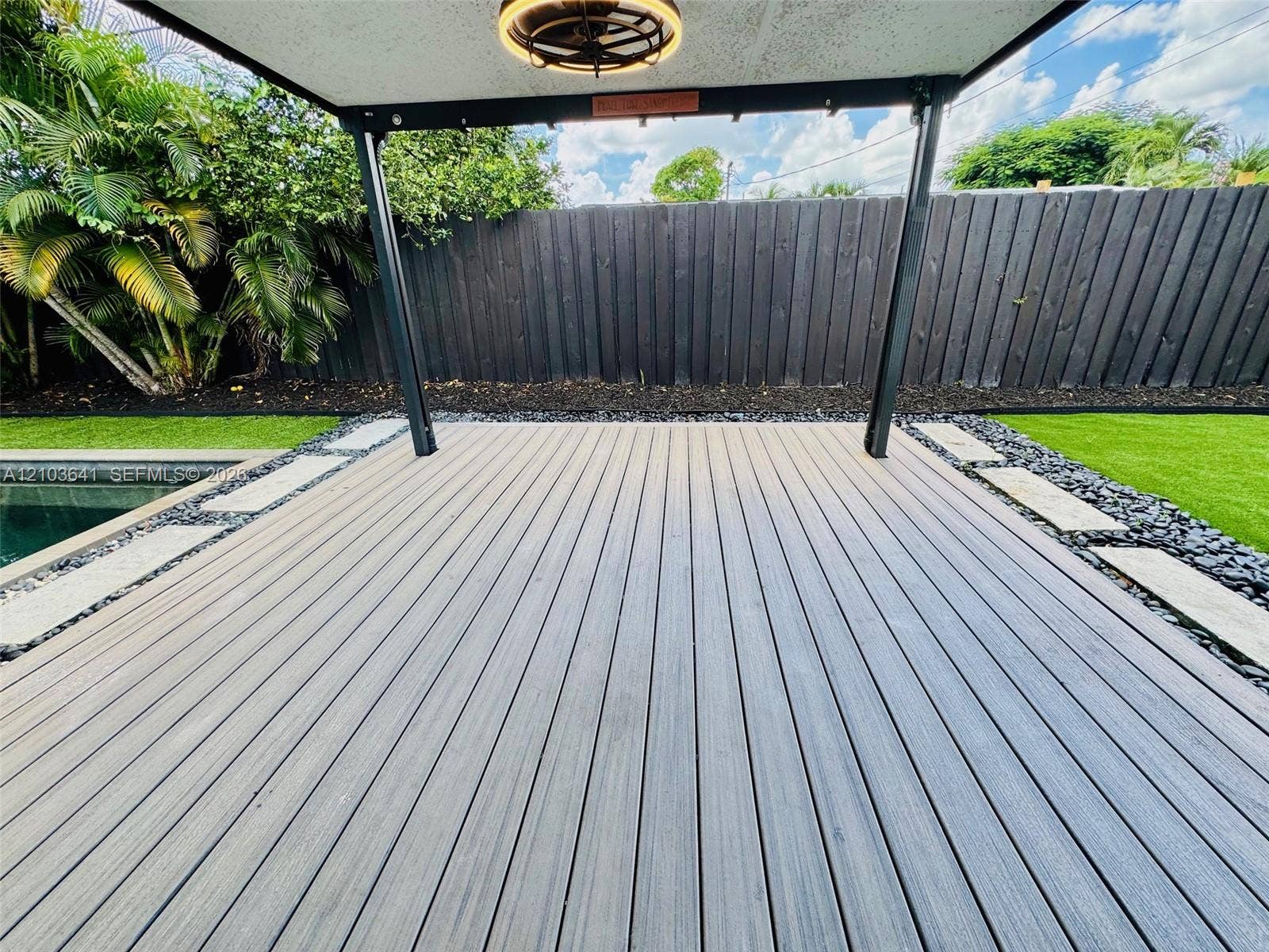 Covered outdoor deck featuring composite decking, a built-in overhead light fixture, stone-lined paver borders, and artificial turf surroundings