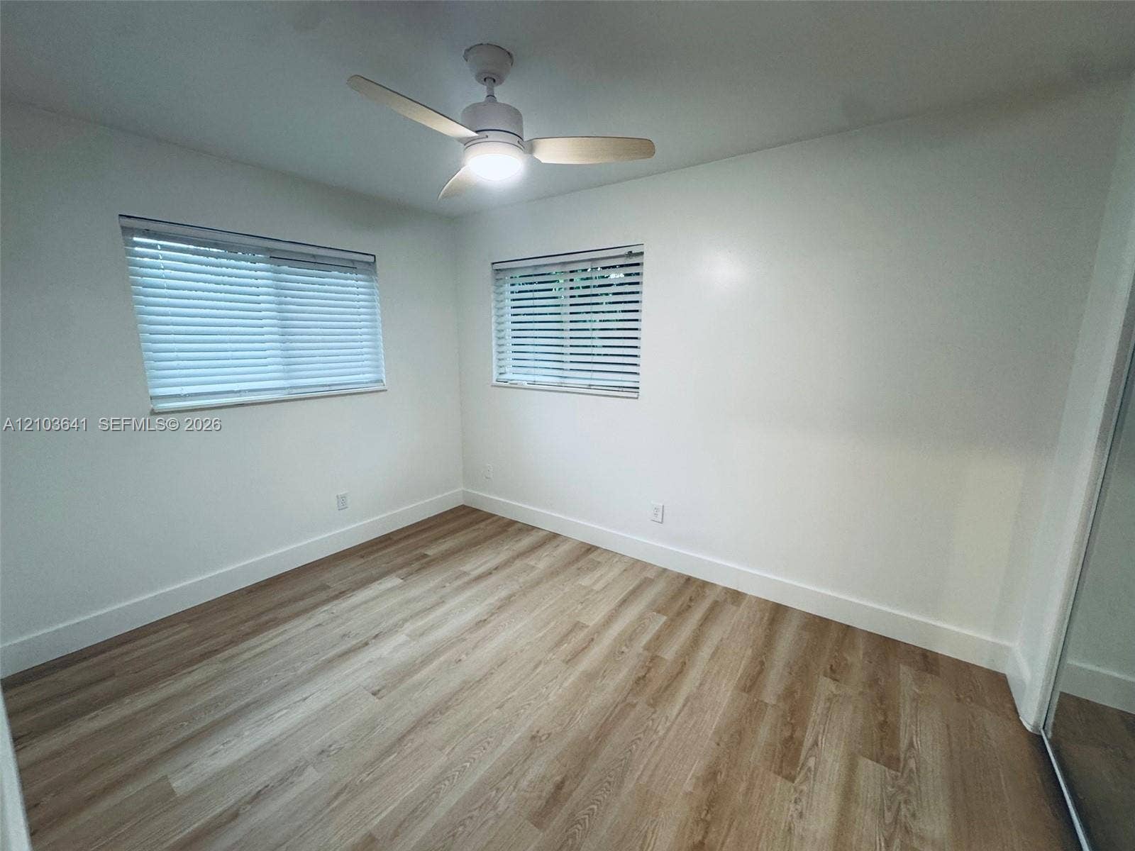 Room featuring wood-finish flooring, dual windows with horizontal blinds, and a white ceiling fan with integrated lighting