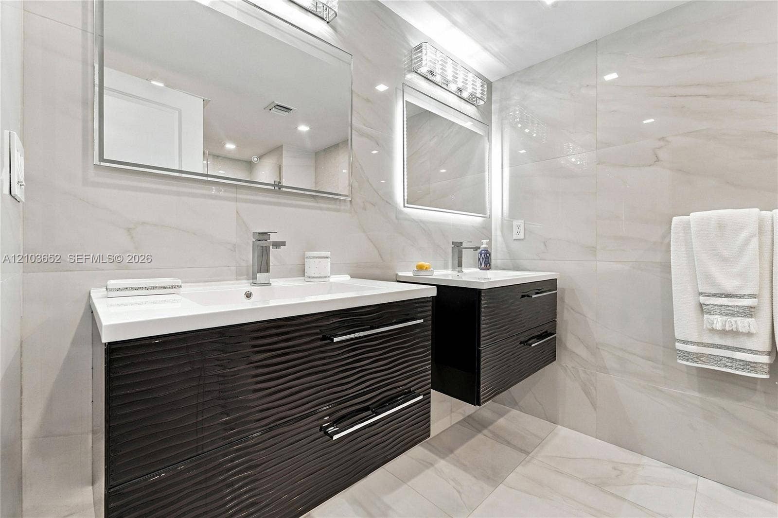 Dual floating vanities featuring dark-toned textured cabinetry, white integrated sinks, polished chrome fixtures, and full-wall porcelain tiling