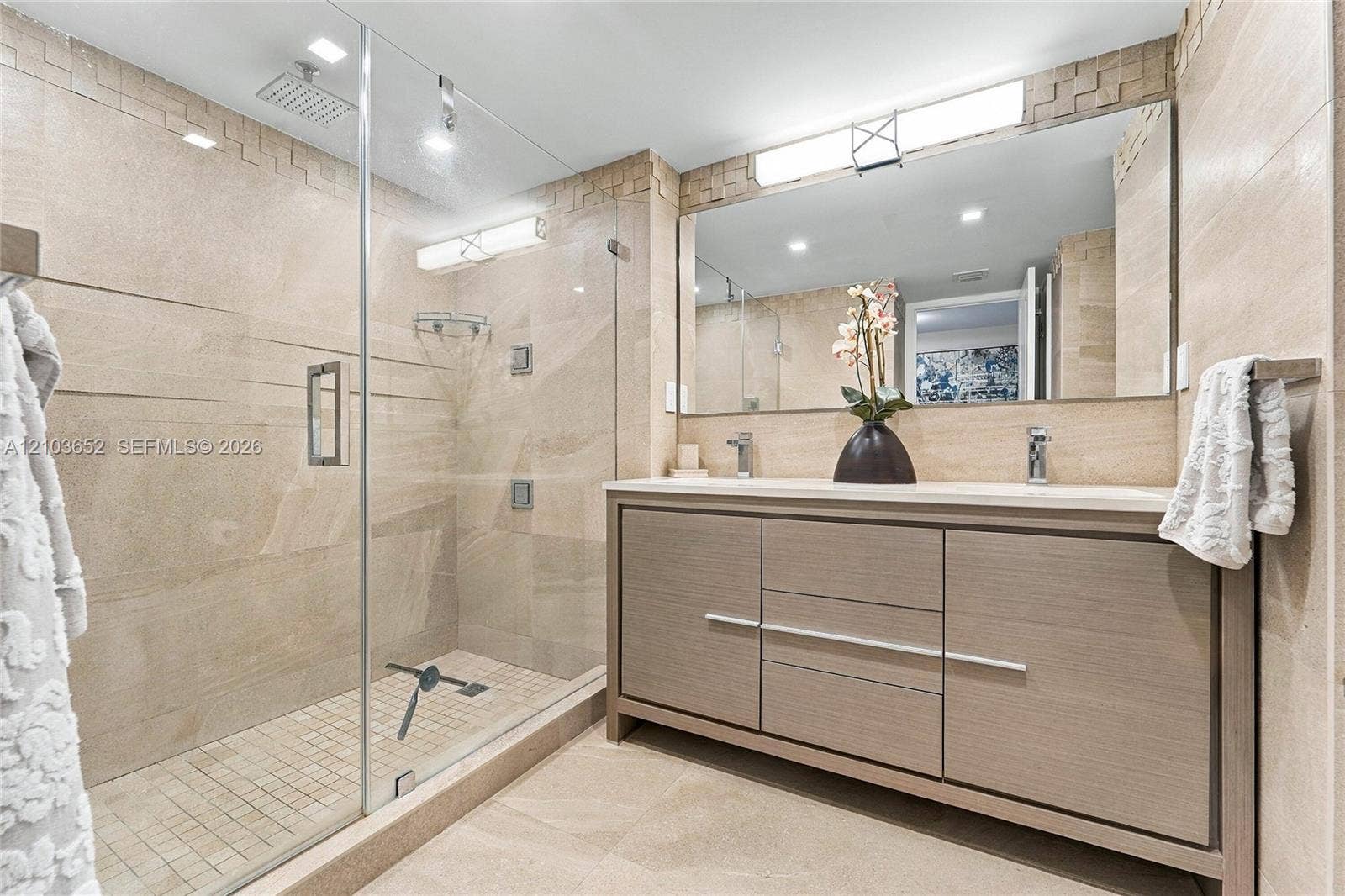 Bathroom featuring a walk-in glass-enclosed shower with a rainfall showerhead, wood-finish vanity with dual integrated sinks, and neutral stone-style wall and floor tiling