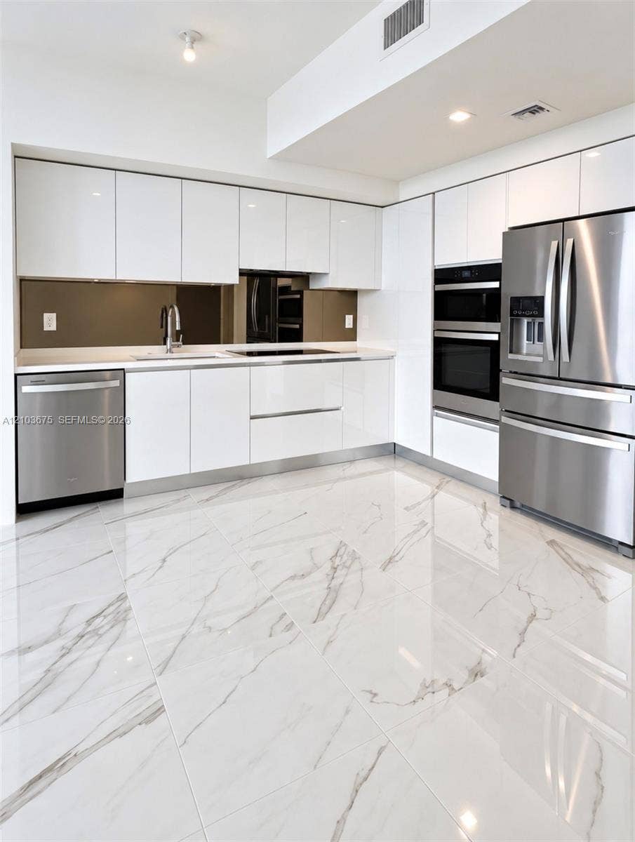 High-gloss flat-panel white cabinetry, stainless steel appliances, integrated double wall ovens, mirror-finish backsplash, and polished stone-finish tile flooring