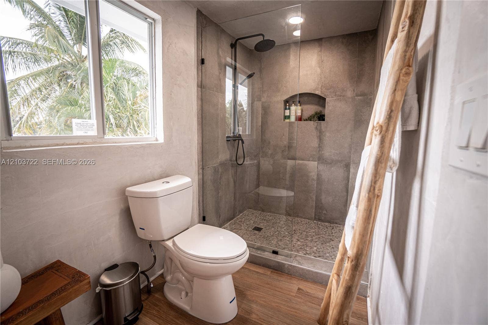 Walk-in shower featuring large-format tile surround, pebble flooring, built-in wall niche, and matte black rainfall shower fixture with handheld attachment, complemented by wood-finish flooring