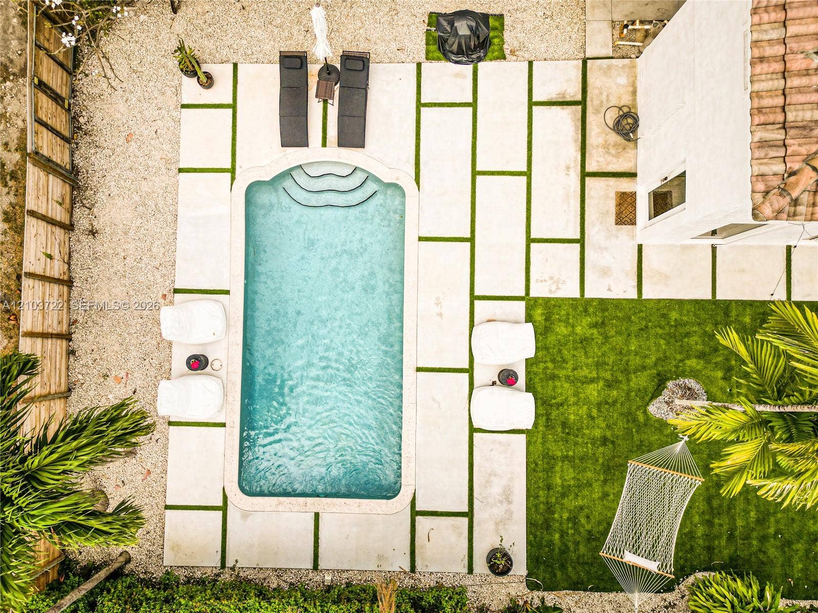 Rectangular swimming pool with built-in steps, light-colored paver decking with artificial turf accents, synthetic lawn area, and wood-style privacy fencing