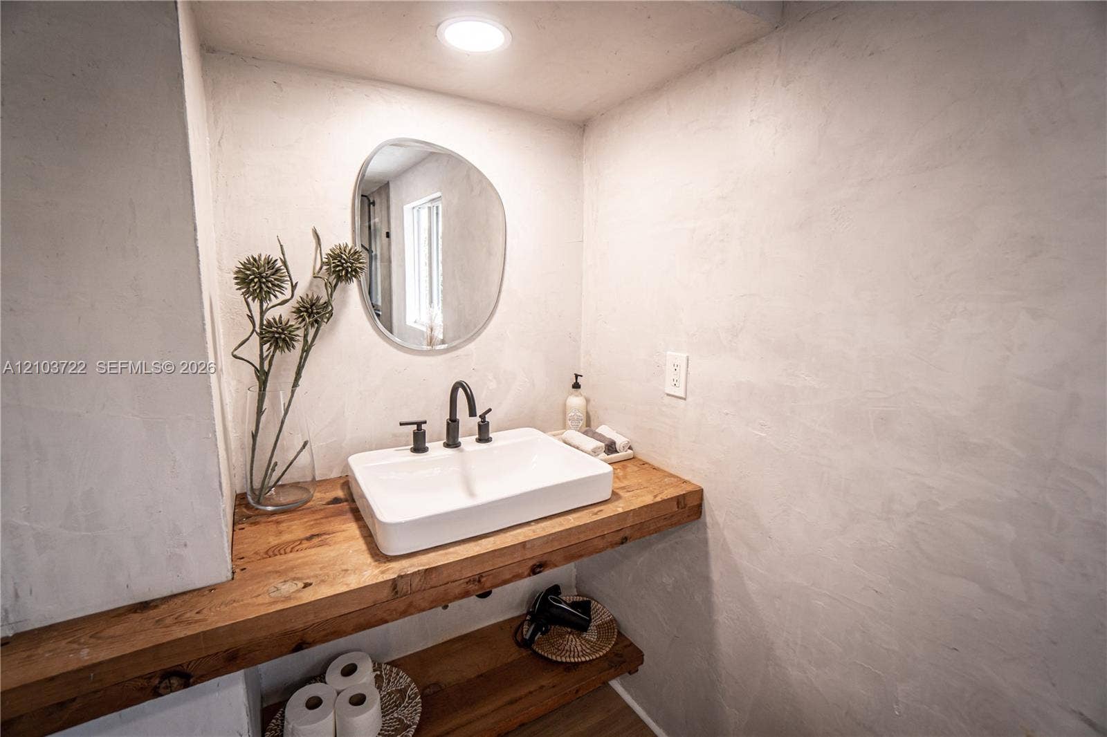 Floating wood-finish vanity with a rectangular vessel sink, matte black widespread faucet, organic-shaped mirror, and recessed ceiling lighting against a textured plaster wall finish