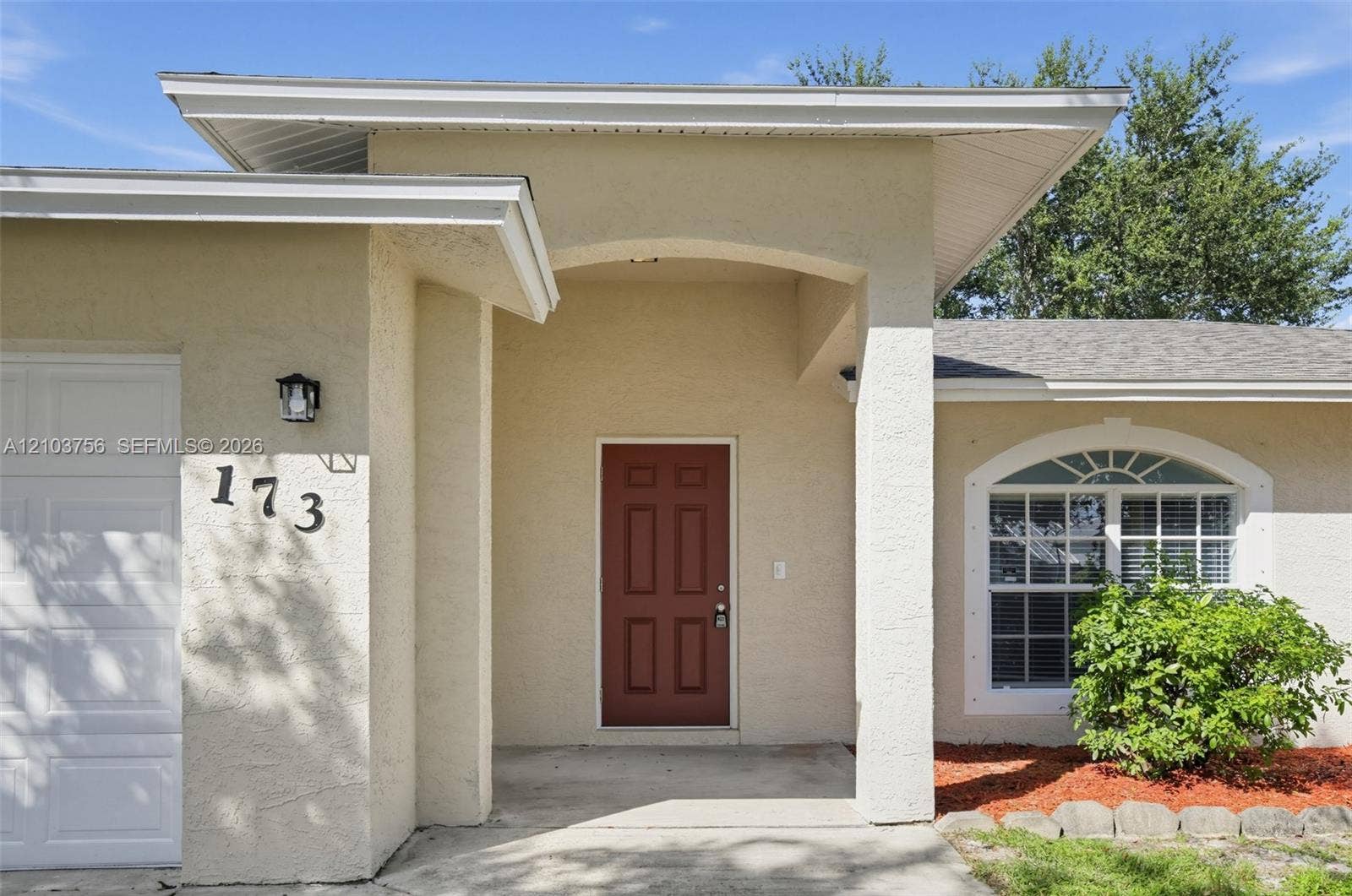 Single-story exterior featuring light-toned stucco siding, an attached two-car garage, an arched entryway, and multiple windows with decorative arched transoms
