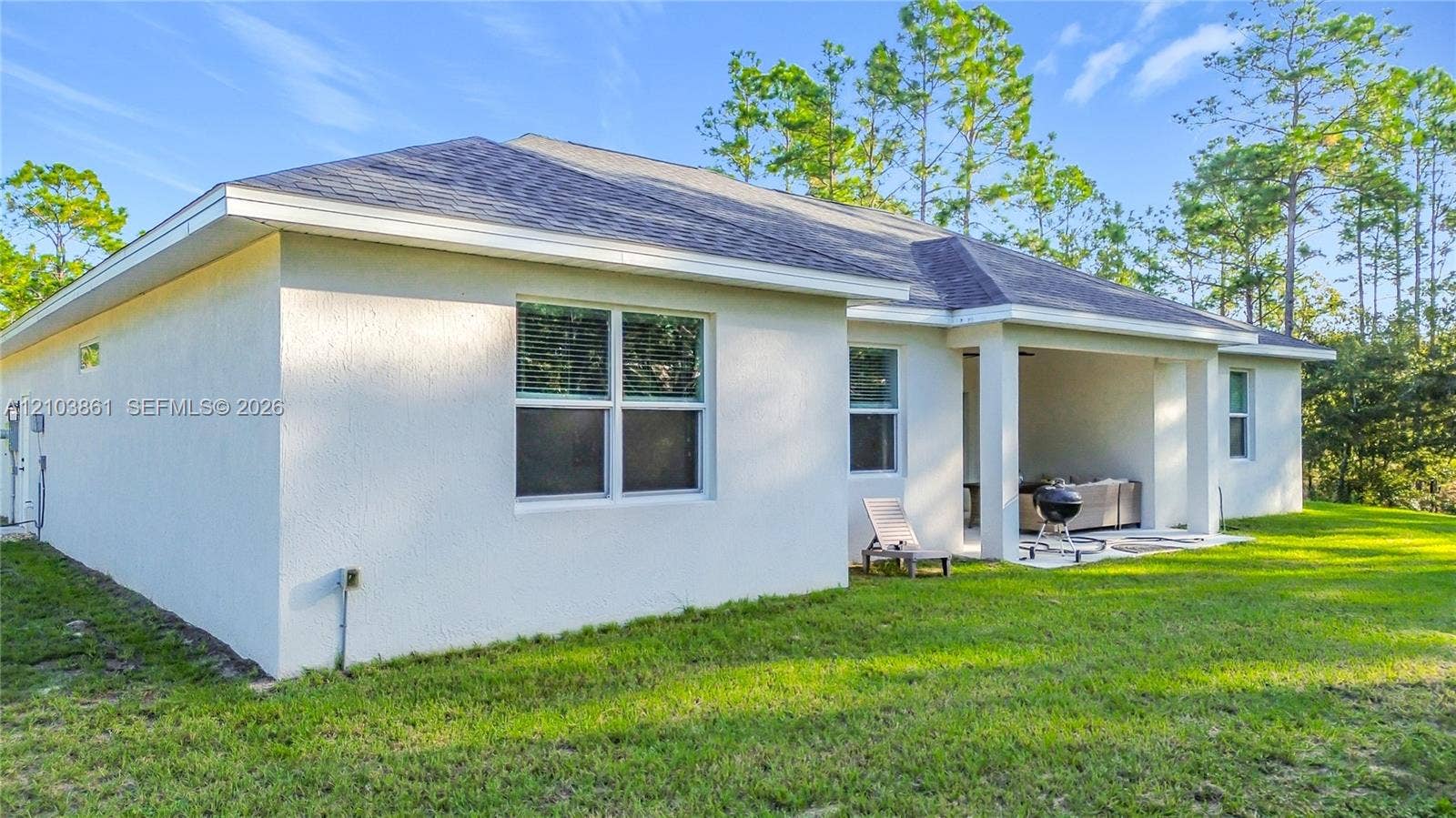 Stucco exterior featuring a covered patio, shingle roof, and expansive backyard with mature pine trees