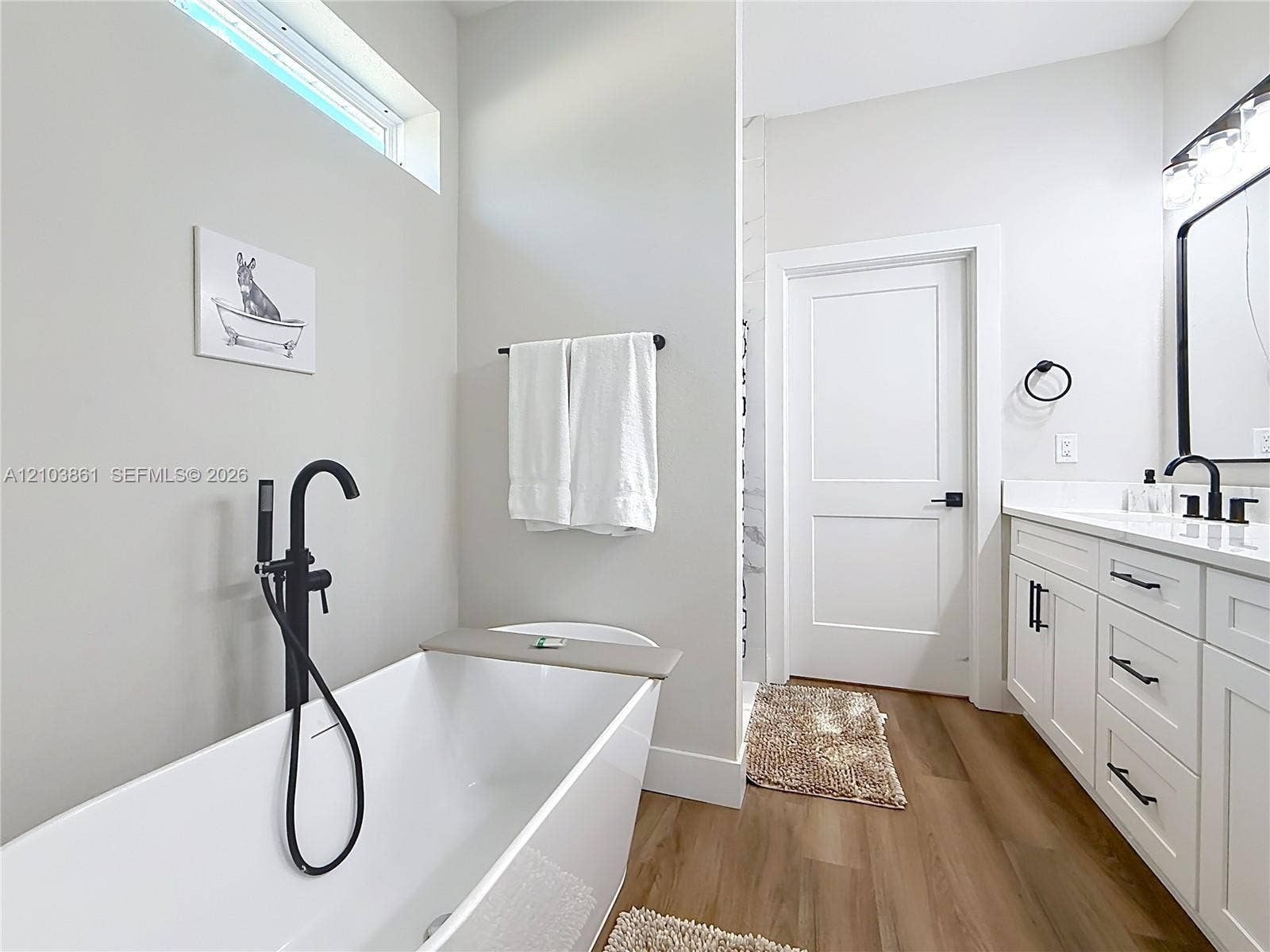 Freestanding soaking tub with a matte black floor-mounted faucet, wood-finish flooring, white shaker-style vanity cabinet with integrated countertop, and transom window