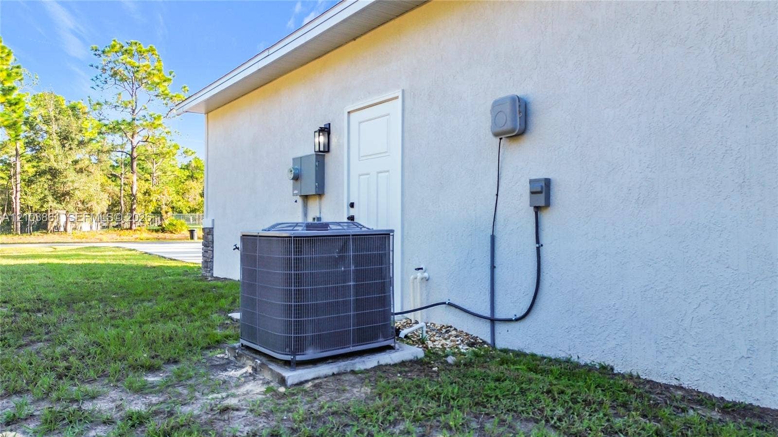 Exterior stucco wall featuring a single-entry door, external wall-mounted lighting, and a central heating and cooling unit on a concrete pad