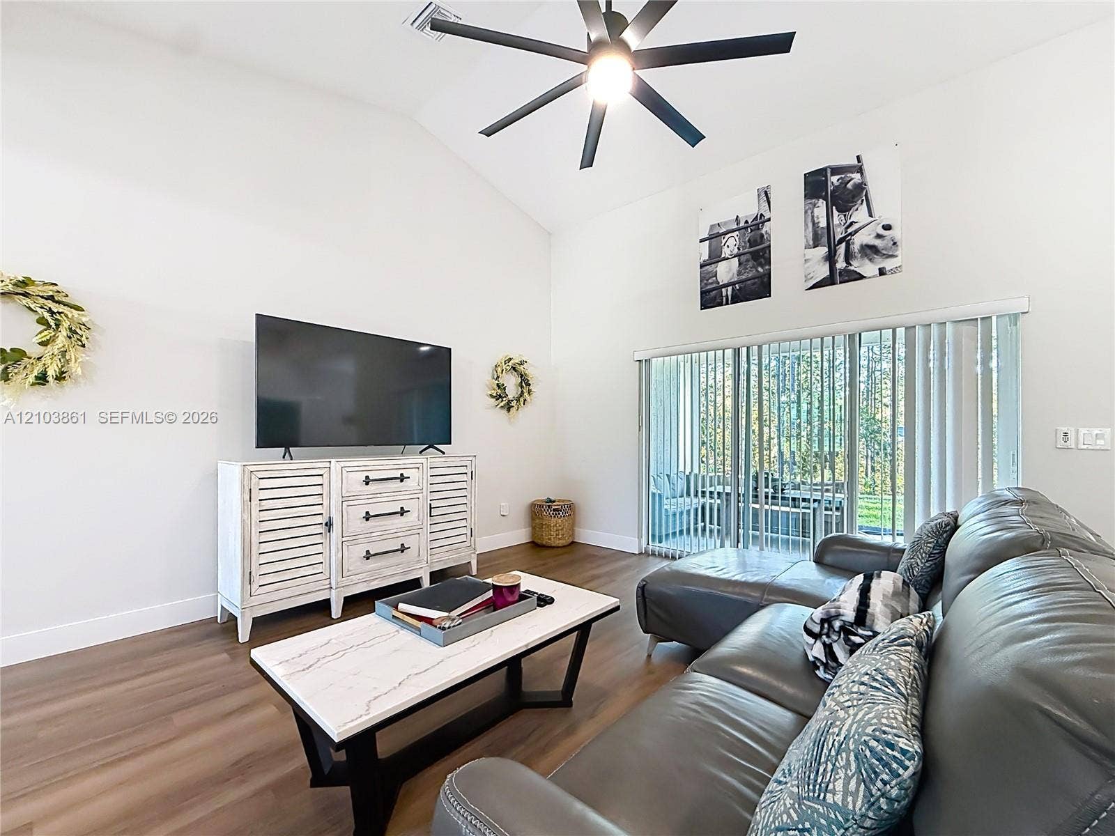 Vaulted ceiling featuring a central matte black ceiling fan, wood-finish flooring, and sliding glass patio doors with vertical blinds