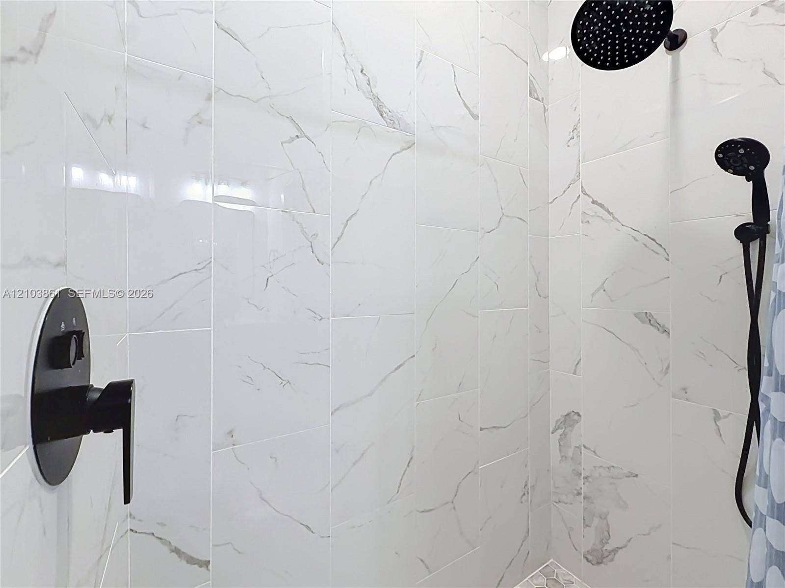 Walk-in shower featuring large-format stone-patterned wall tile, matte black rainfall showerhead, adjustable handheld shower wand, and a matching matte black pressure-balance faucet