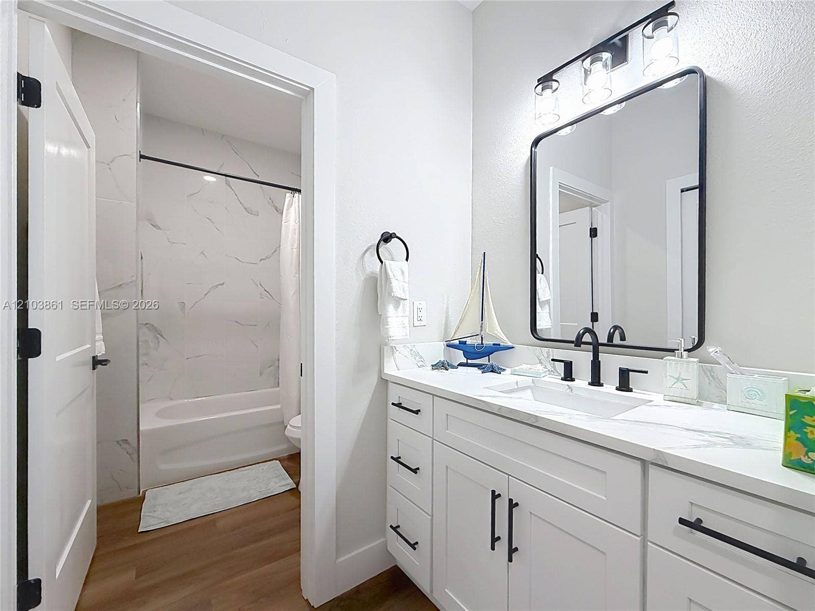 Bathroom featuring a white vanity with stone-style countertop, rectangular undermount sink, matte black hardware, framed vanity mirror, and tub-shower combination with floor-to-ceiling tile surround