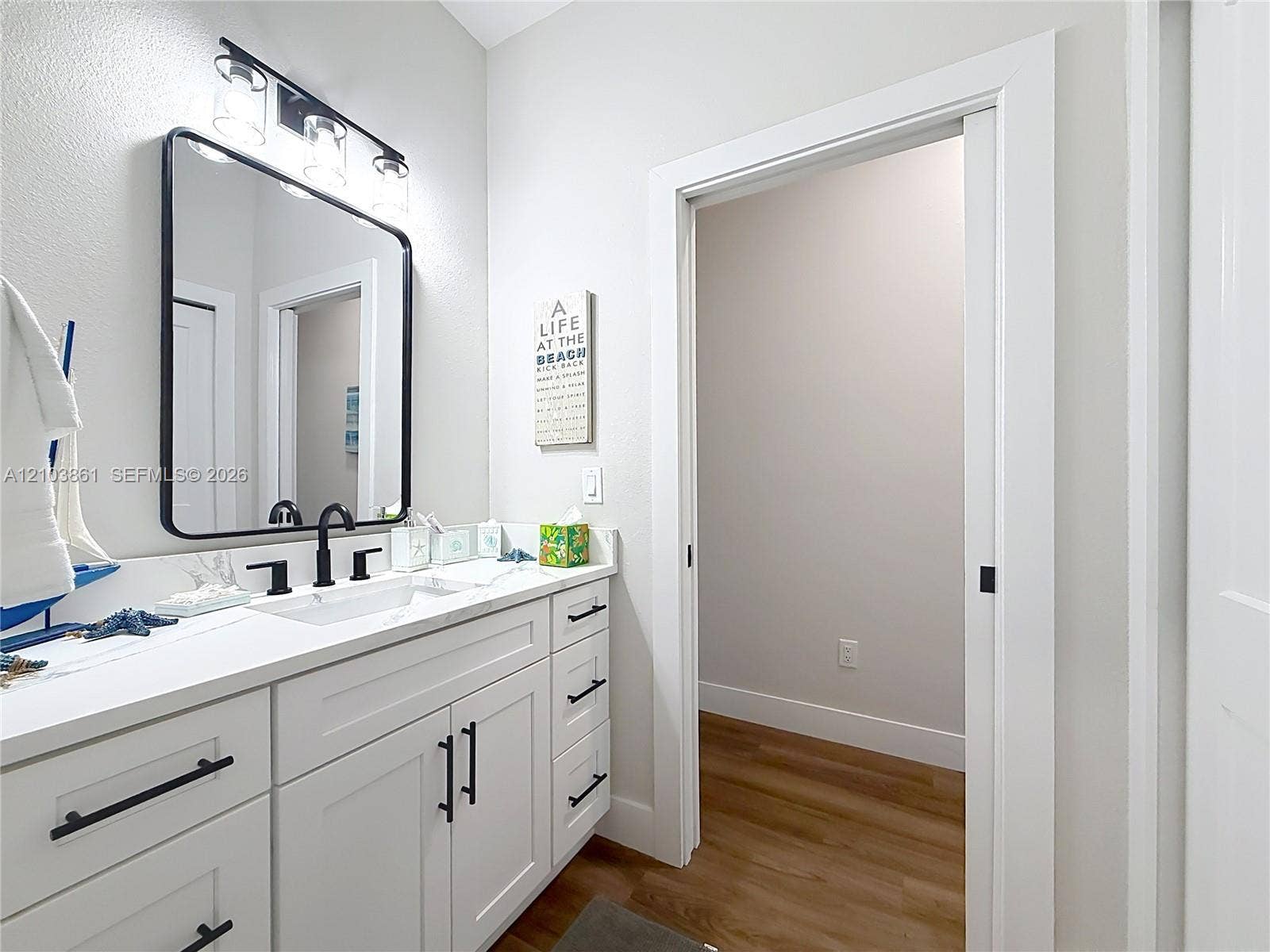 White cabinetry vanity with stone-finish countertop, undermount sink, matte black hardware, rectangular framed mirror, and wood-finish flooring