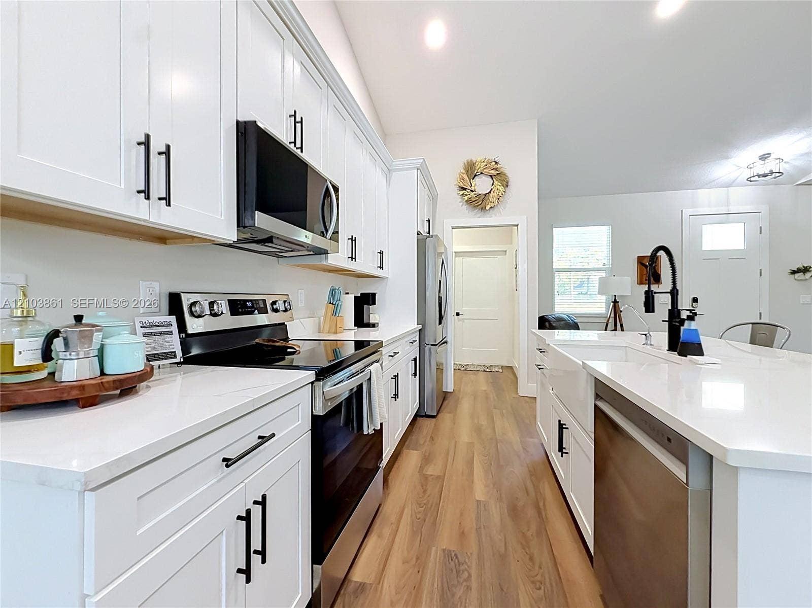 Kitchen featuring white shaker-style cabinetry, quartz countertops, stainless steel appliances, a farmhouse-style apron sink with a matte black faucet, and wood-finish flooring