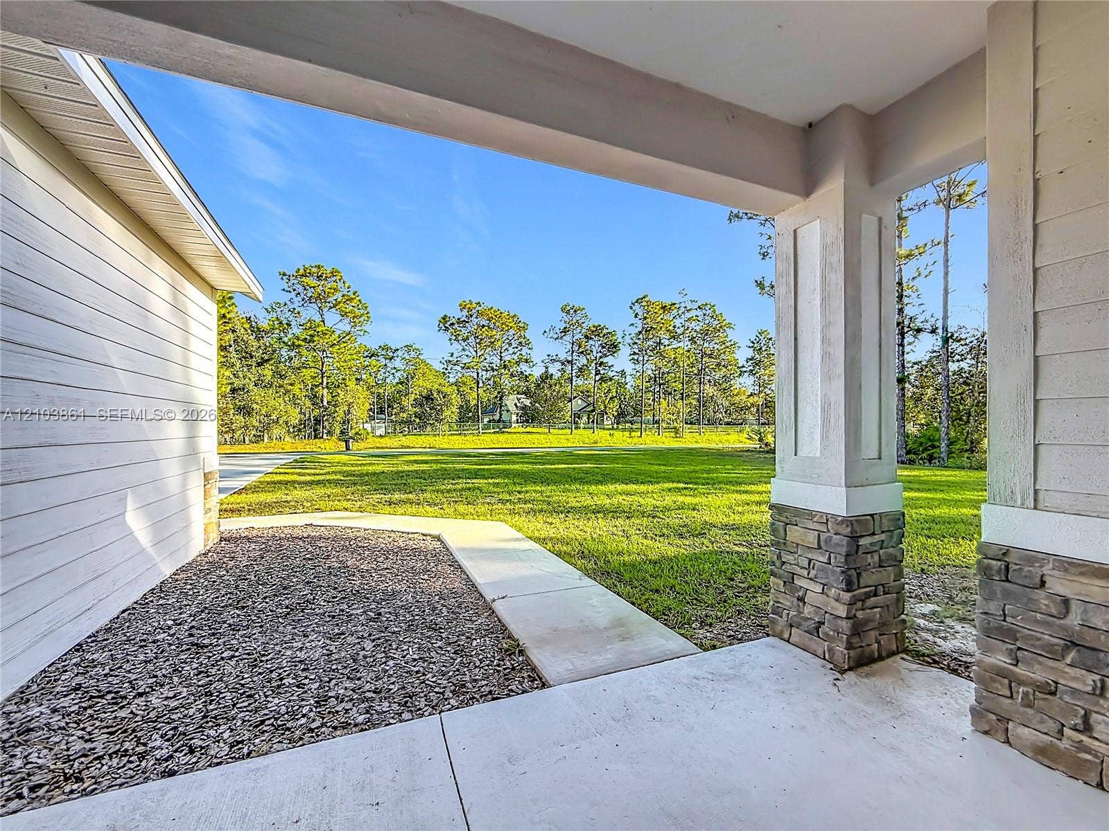 Covered patio featuring concrete flooring, stone-veneer column bases, and horizontal lap siding, opening to a grass-covered backyard with mature trees