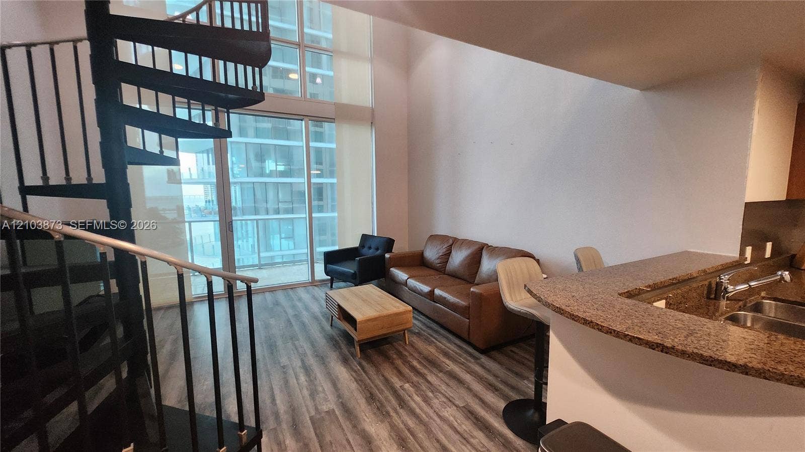 300 S Biscayne Boulevard, #L-808, Miami, FL 33131 Double-height living area featuring floor-to-ceiling windows, a black metal spiral staircase, wood-finish flooring, and a kitchen peninsula with stone-finish countertops