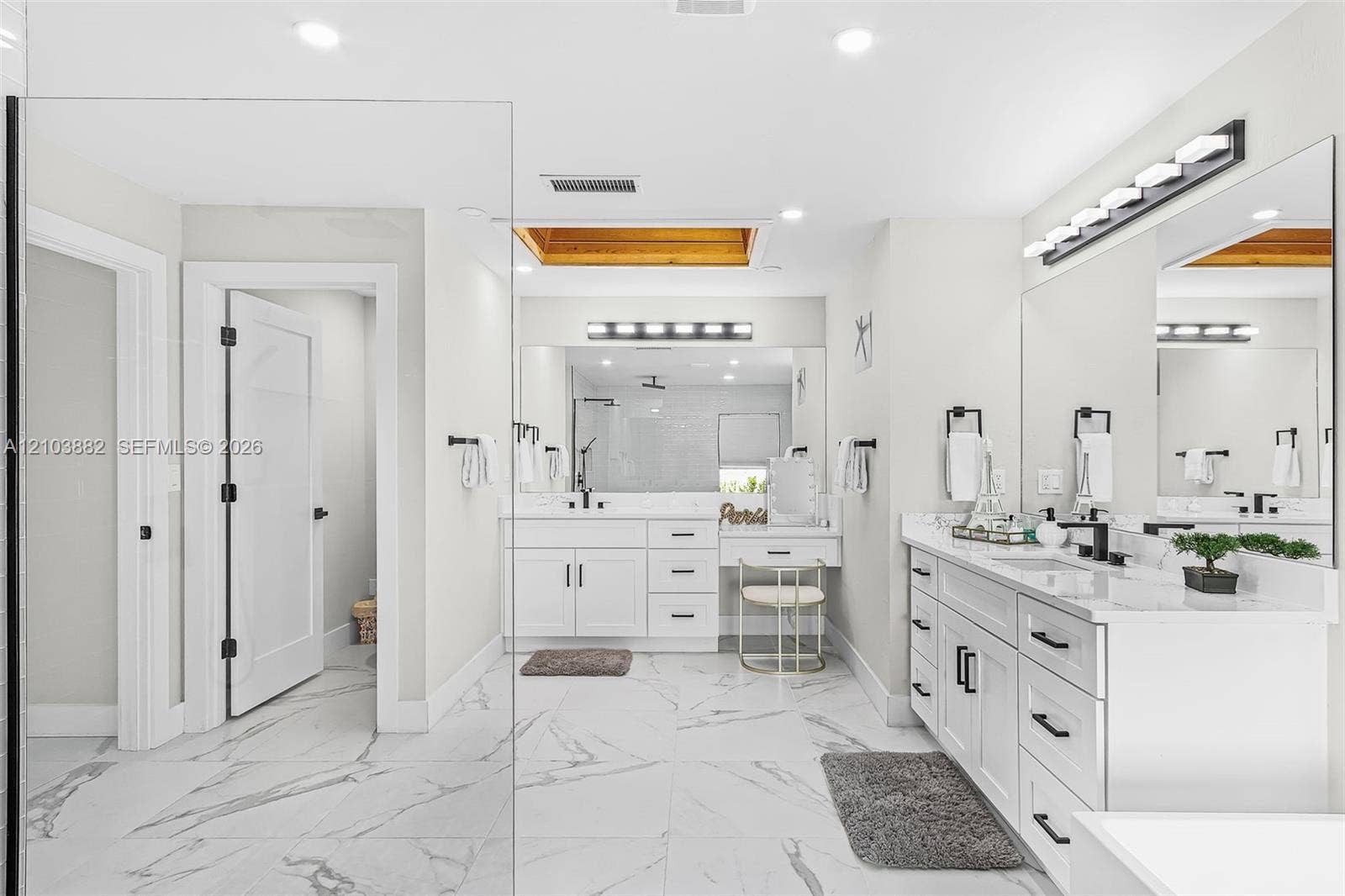 Dual white vanities with light-toned stone countertops, large-format tile flooring, recessed skylight, and wall-mounted linear vanity lighting