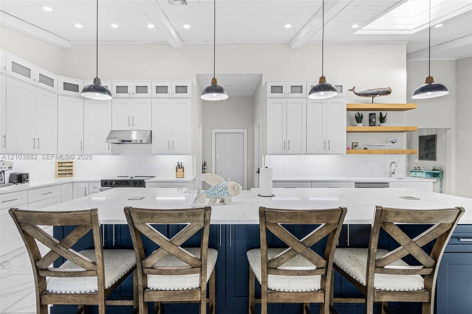 Kitchen featuring white upper cabinetry, a navy-blue central island with a stone countertop, floating wood shelves, stainless steel appliances, and pendant lighting