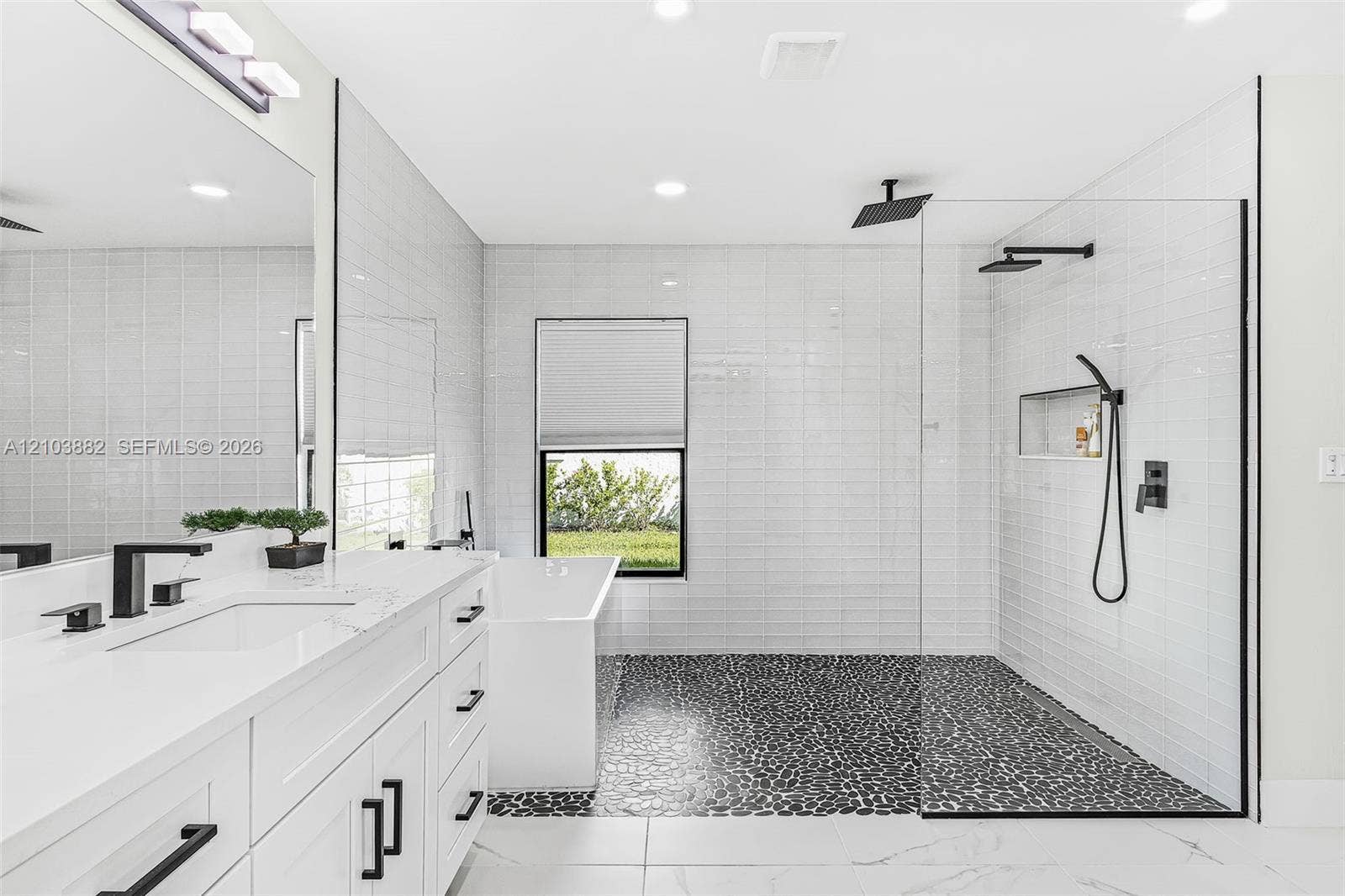 White vanity with stone countertop, freestanding bathtub, walk-in shower with river stone flooring, glass enclosure, and matte black fixtures