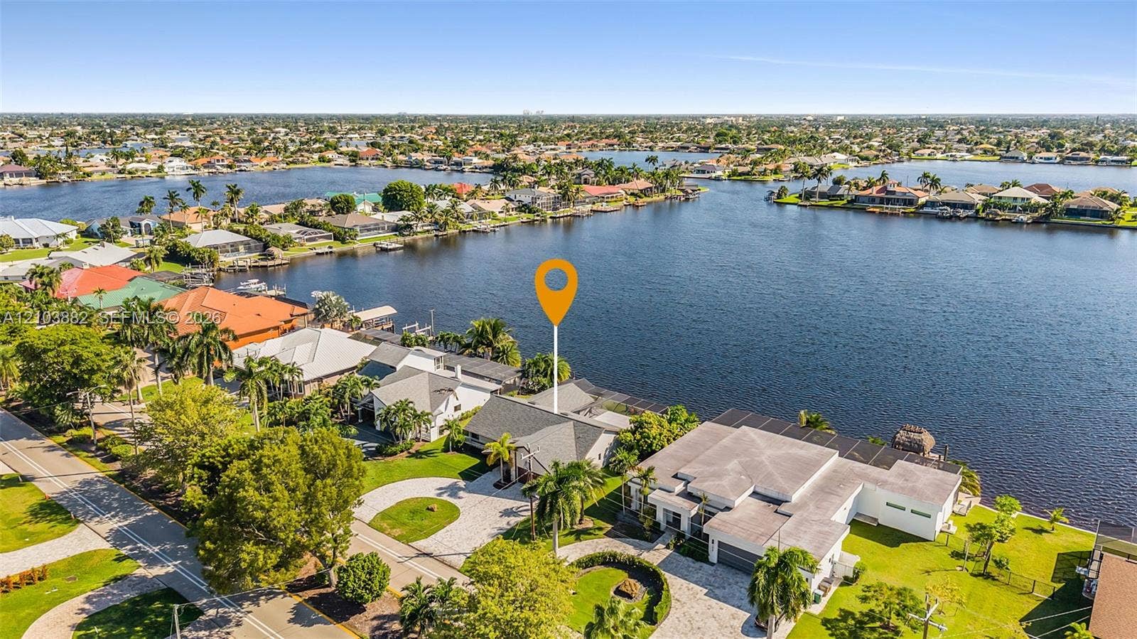 Aerial perspective featuring expansive water frontage, paved circular driveway, mature palms, and residential roofline