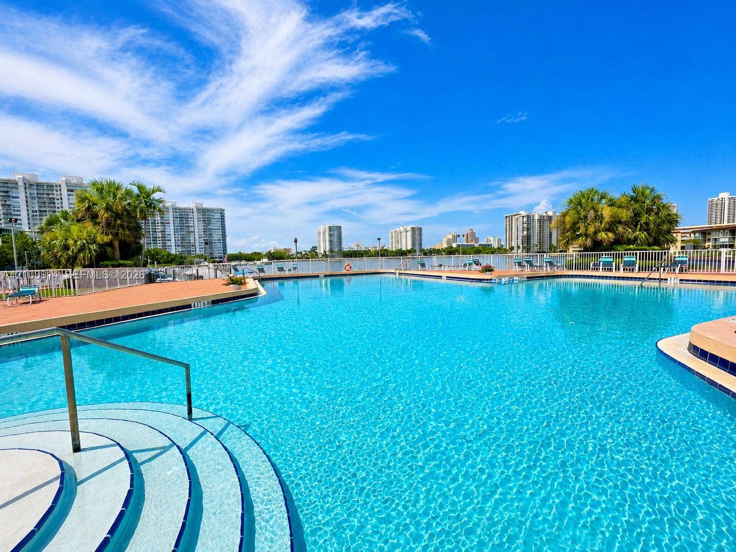 Swimming pool featuring submerged entry steps, a metal handrail, paved pool deck, and perimeter fencing