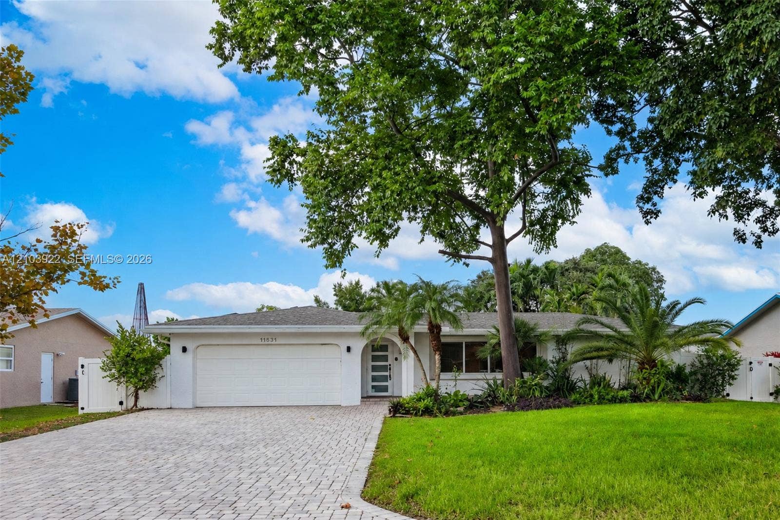 White-painted exterior with a paver driveway, an attached garage, mature trees, and established landscaping