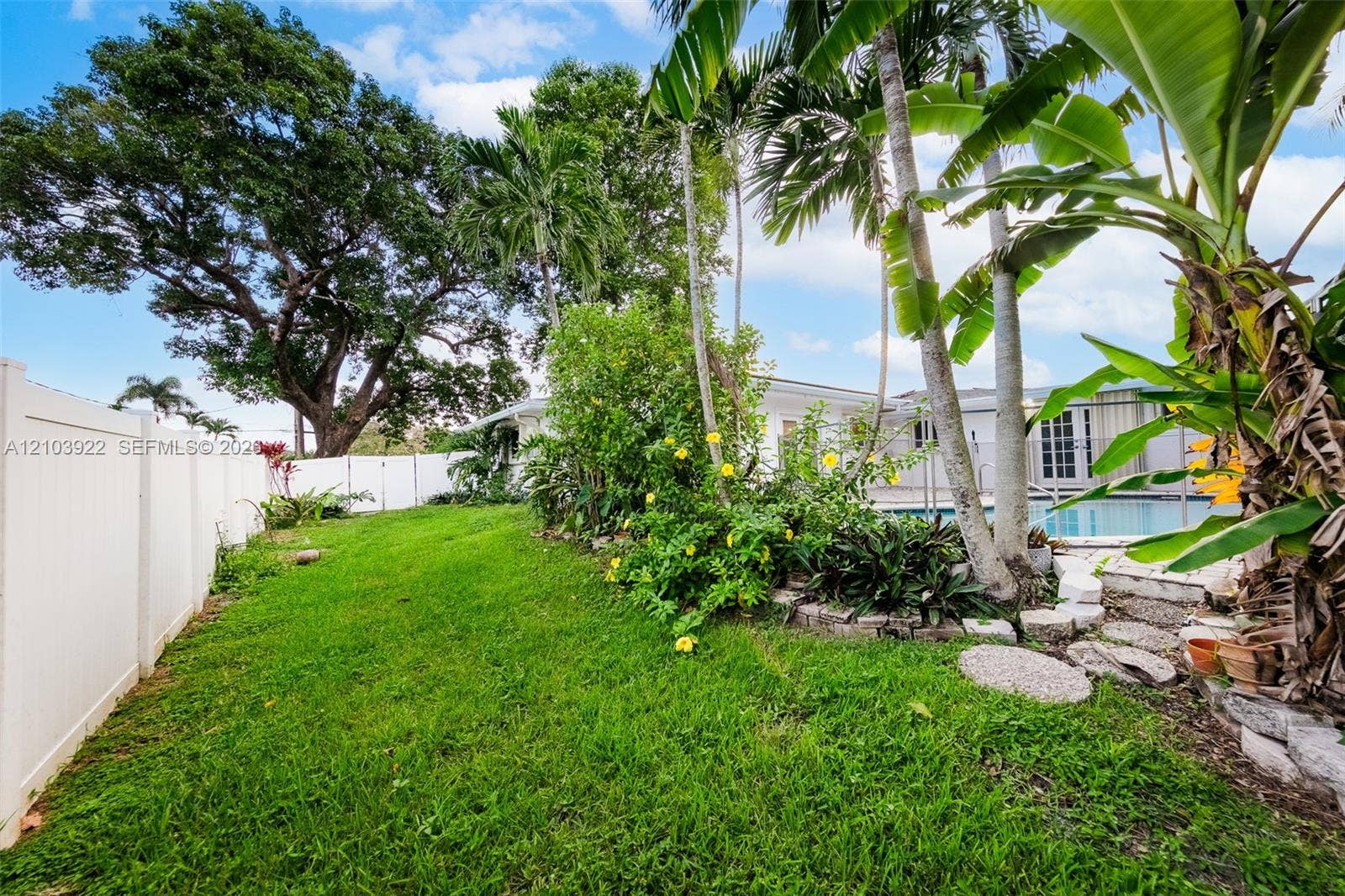 Expansive rear yard featuring green lawn, white privacy fencing, mature trees, tropical foliage, and a visible swimming pool