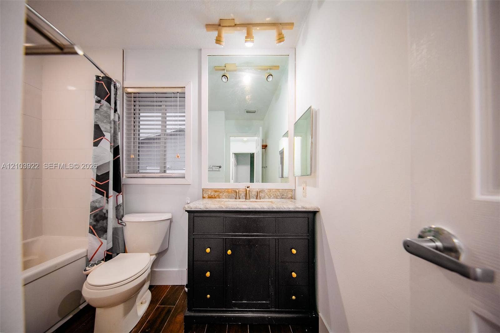 Dark wood-finish flooring, built-in vanity with a light-colored countertop, wall-mounted mirror, and a window with blinds