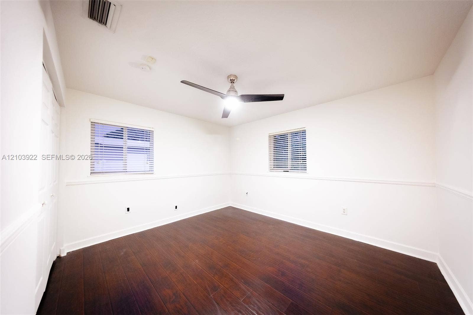 Spacious room featuring rich wood-finish flooring, two windows with horizontal blinds, a modern ceiling fan, and crisp white walls with wainscoting