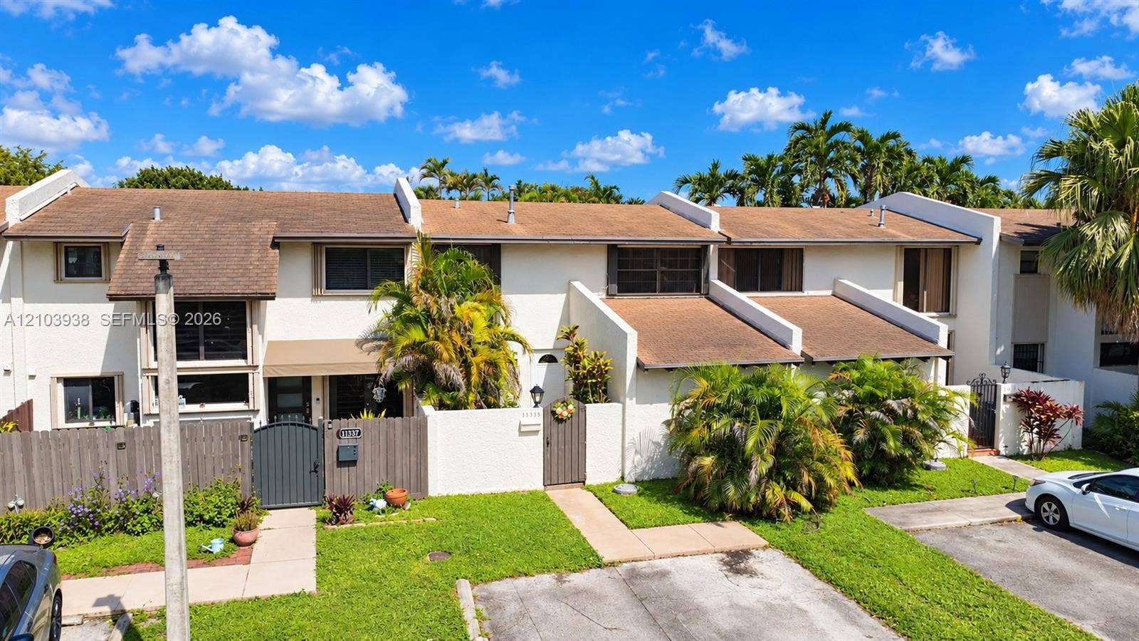 11335 SW 71st Lane, Miami, FL 33173 Townhome exterior featuring light-colored stucco siding, a multi-level shingle roofline, private courtyard entryways with metal gates, and dedicated paved parking