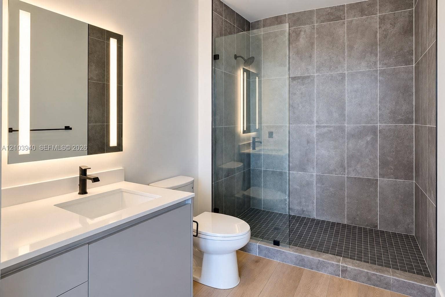307 SW 5th Street, #2610, Fort Lauderdale, Fort Lauderdale, FL 33315 Bathroom featuring a vanity with a white countertop, wall-mounted mirror with integrated lighting, glass-enclosed walk-in shower with grey wall tile, wood-finish flooring, and matte black hardware